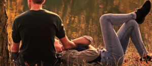 Image of a couple sitting outdoors facing away from the camera, with one partner sitting crosslegged and the other partner lying back on the ground with their head in the other person's lap.