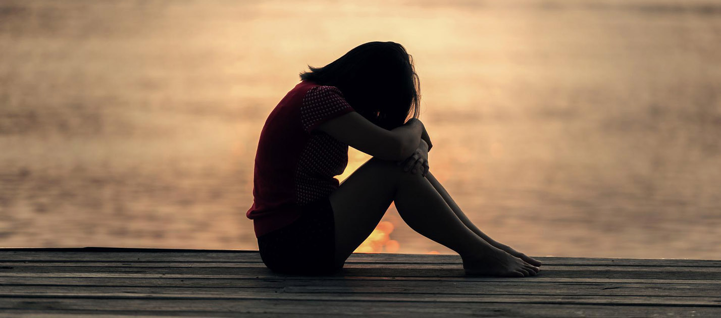 Silhouette of young person sitting on a dock with their knees pulled up and head bowed