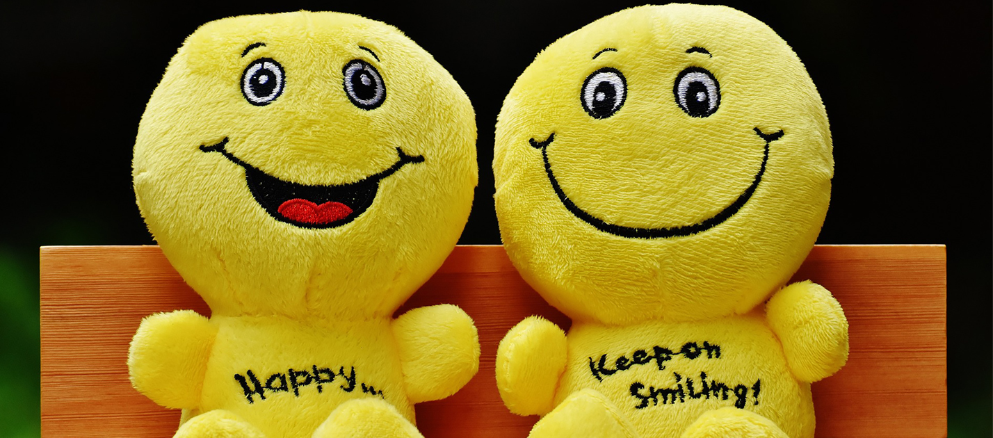 Two yellow happy face stuffies sitting side by side on a bench. One stuffie's belly says "Happy" and the other's says "Keep Smiling"