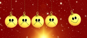 A row of four yellow holiday ornaments with frowny faces hangs against a red background with sparkles of light, as a fifth yellow ornament with a smiley face swings into the others like a Newton's Cradle.